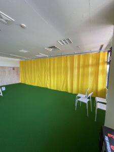 愛知県北名古屋市のスポーツ教室で行った仕切りカーテン設置工事。防炎機能付きカーテンのターポリン イエロー生地を使用し、フロアを2つに仕切って空間を有効活用。集中できる環境づくりや安全性の向上を目的とした施工。